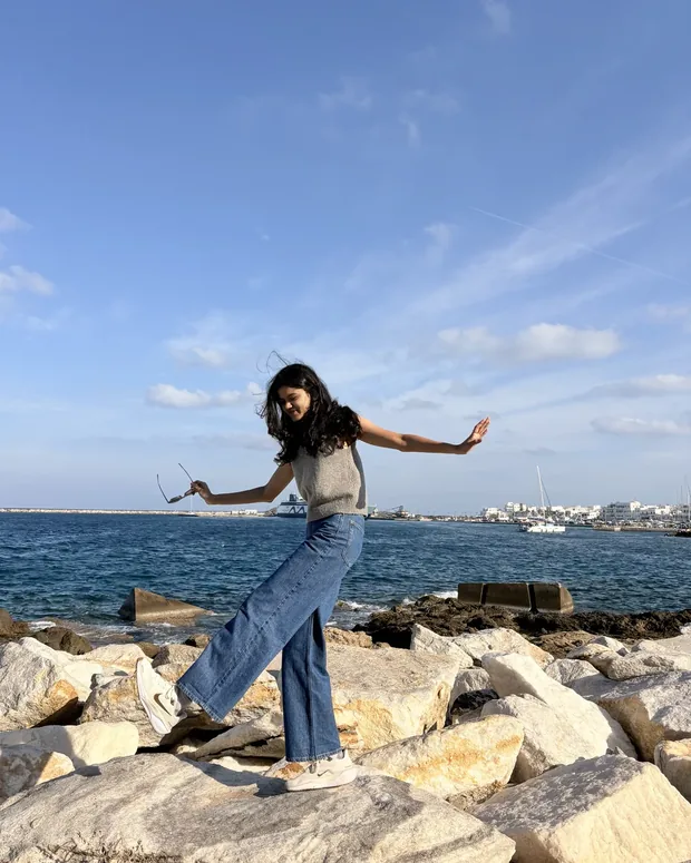 Gauri mid-stride across the rocks at the water's edge, sunglasses in one hand and the other out for balance, a harbour and open sea behind her
