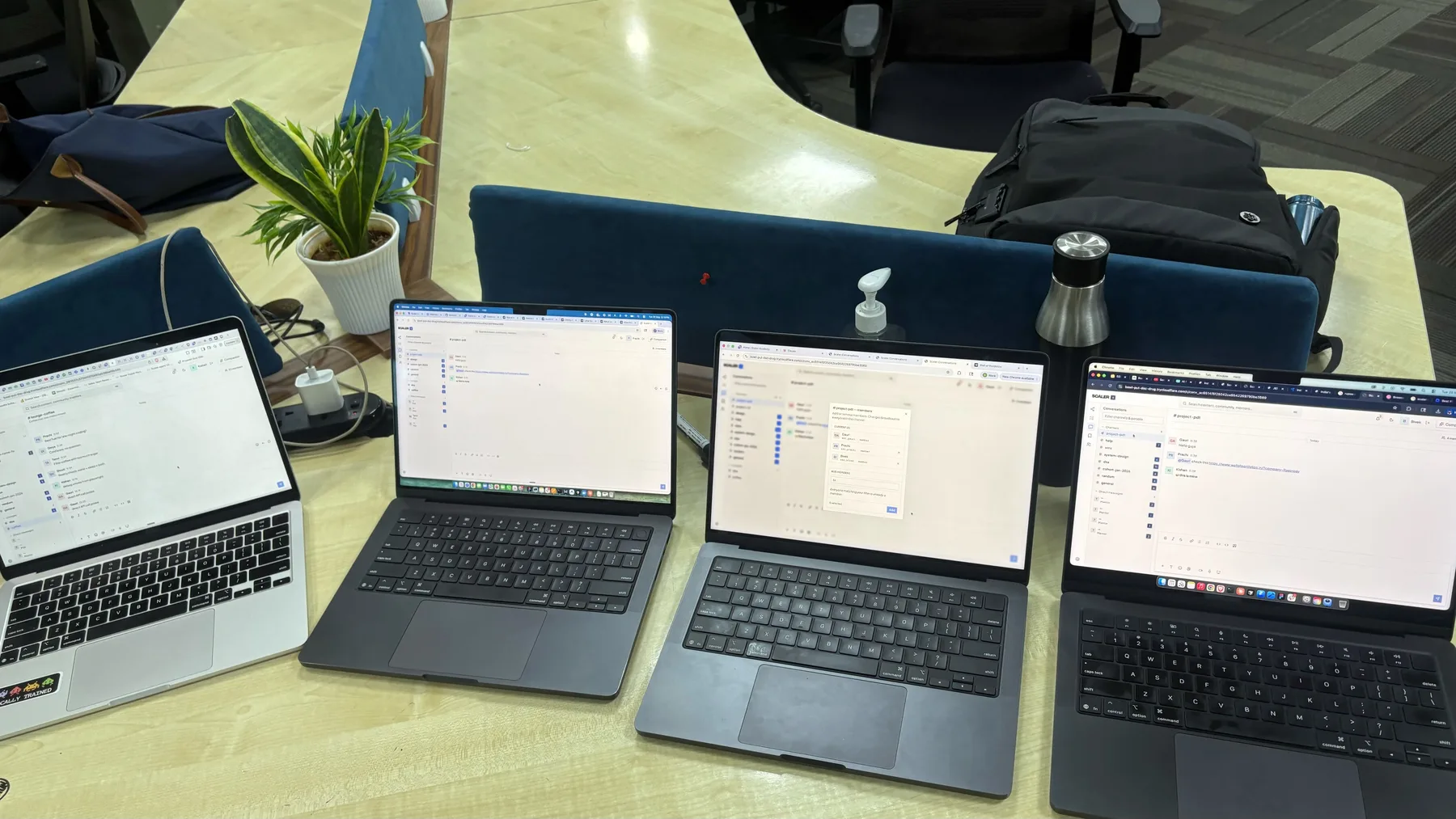 Four laptops open side by side on an office table, each signed into a different account in the same running build: two showing the #project-pdt channel, one with the add-members panel open on the channel roster, one with a mention in the thread.